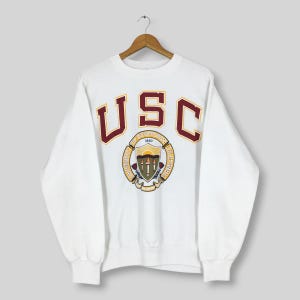 Puede incluir: Sudadera blanca de cuello redondo con las letras "USC" en granate y dorado. La sudadera presenta un escudo universitario en el centro. Cuelga de una percha de madera.