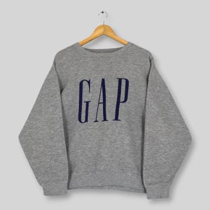 May include: A heather grey long-sleeve crewneck sweatshirt featuring the word GAP printed in dark navy blue capital letters across the chest. The garment is displayed on a wooden hanger, showcasing a ribbed neckline, cuffs, and hem.