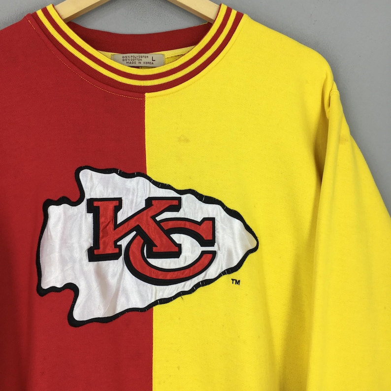 May include: A two-toned, long-sleeved sweatshirt. The left side is red, and the right side is yellow. The front features a white and red logo with the letters "KC" inside a white arrowhead. The collar is yellow with red and yellow stripes.