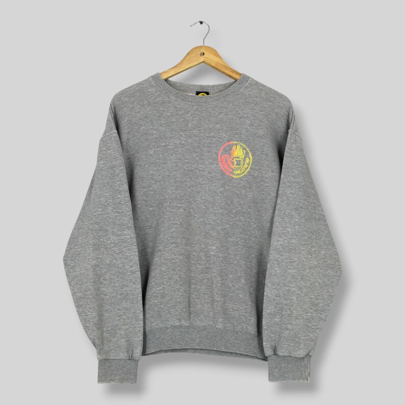May include: A gray sweatshirt with a crew neck and long sleeves. A circular graphic with red, yellow, and orange hues is centered on the chest. The sweatshirt is hanging on a wooden hanger.