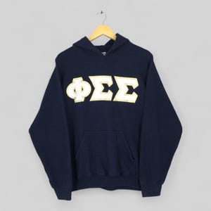 May include: Navy blue hooded sweatshirt with a large white and gold Greek letter design across the chest. The sweatshirt has a front pocket and a drawstring hood. The garment is hanging on a wooden hanger.