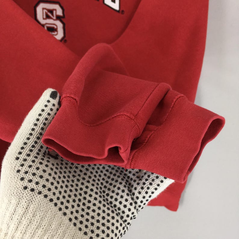 May include: A red long-sleeve sweatshirt featuring a ribbed cuff at the wrist. The fabric is a solid red color. A partial graphic is visible at the top of the garment, displaying the letters NC in a white block font with a black outline.