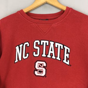 May include: A red crewneck sweatshirt featuring the text NC STATE in white letters with black outlines arched across the chest. Below the text is a white and black block letter S with the letters NC inside. The garment has a ribbed collar.