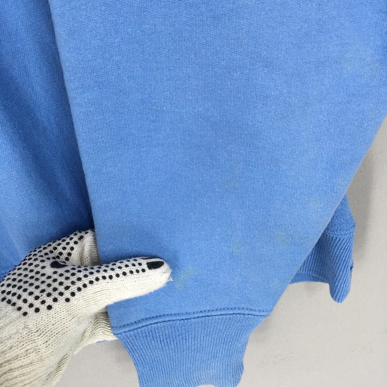 May include: A pale blue sweatshirt with a matching ribbed cuff and hem. The sleeve is being held by a gloved hand. There are some stains on the sleeve.