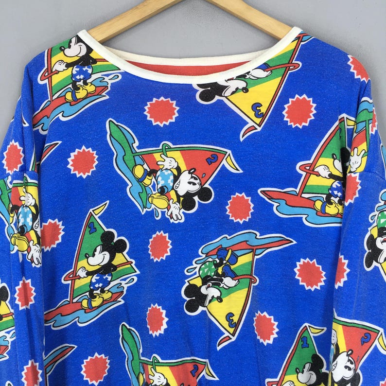 May include: A blue long-sleeve shirt featuring an all-over print of Mickey Mouse windsurfing on colorful sails. The design includes red sunburst shapes and white waves against a bright blue background, finished with a cream-colored crew neckline.