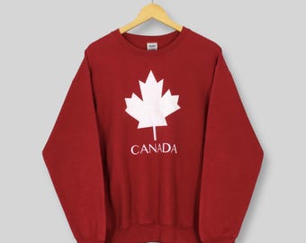 May include: A maroon sweatshirt with a white maple leaf graphic and the word "CANADA" printed below it. The sweatshirt is hanging on a wooden hanger against a light gray background.