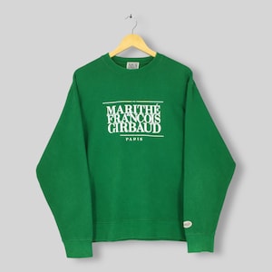 May include: A green sweatshirt featuring the white text "MARITHÉ FRANÇOIS GIRBAUD PARIS". The sweatshirt is suspended from a wooden hanger. The garment has long sleeves and a crew neck. The background is a light grey colour.