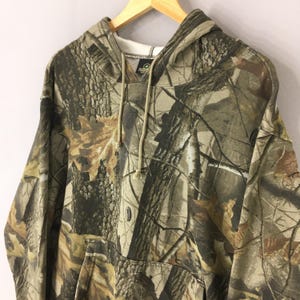 Peut inclure: Un sweat &agrave; capuche camouflage avec une capuche &agrave; cordon et une poche kangourou. Le motif pr&eacute;sente des nuances de marron, de vert et de beige, ressemblant &agrave; un environnement forestier. Le sweat est suspendu &agrave; un cintre en bois.