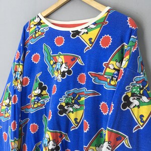 May include: A long-sleeve blue sweatshirt featuring an all-over print of Mickey Mouse windsurfing on colorful triangular sails. The design includes small red sunburst shapes scattered across the fabric and a cream-colored ribbed crew neckline.