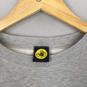 May include: Close-up of a gray crew neck sweatshirt with a black and yellow Body Glove logo patch. The logo features a yellow hand with the words "BODY GLOVE" in a circular design. The sweatshirt is hanging on a wooden hanger.