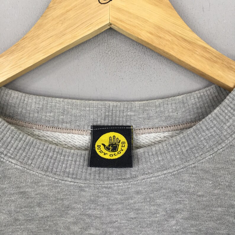 May include: Close-up of a gray crew neck sweatshirt with a black and yellow Body Glove logo patch. The logo features a yellow hand with the words "BODY GLOVE" in a circular design. The sweatshirt is hanging on a wooden hanger.