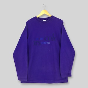 Puede incluir: Una sudadera morada de manga larga con cuello redondo y puños acanalados. El pecho presenta un texto bordado en azul oscuro y negro que dice "colors" y "of benetton" con un pequeño icono de un cuadrado azul sobre la palabra colors.