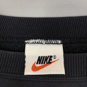 Op de afbeelding: Close-up van een zwart sweatshirt met een wit label met het Nike-logo. Het logo is een oranje swoosh boven het woord "NIKE" in zwarte hoofdletters. Het label is rechthoekig en op het sweatshirt genaaid.