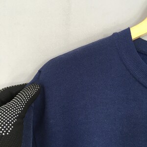 May include: Close-up of a navy blue sweatshirt. The image shows the shoulder and neckline of the garment, with a portion of a gloved hand visible. The sweatshirt is hanging on a wooden hanger against a light grey background.