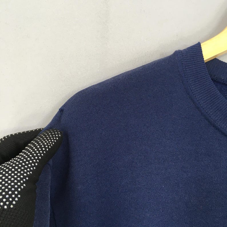 May include: Close-up of a navy blue sweatshirt. The image shows the shoulder and neckline of the garment, with a portion of a gloved hand visible. The sweatshirt is hanging on a wooden hanger against a light grey background.