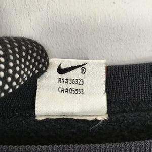 Op de afbeelding: Close-up van een wit label met het Nike swoosh-logo en de tekst "RN#56323" en "CA#05553" in zwart gedrukt. Het label is bevestigd aan een donkergrijze stof.