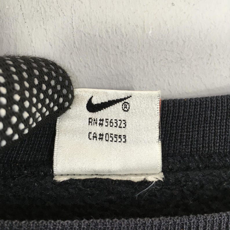 Op de afbeelding: Close-up van een wit label met het Nike swoosh-logo en de tekst "RN#56323" en "CA#05553" in zwart gedrukt. Het label is bevestigd aan een donkergrijze stof.