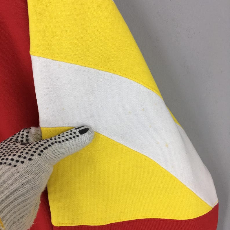 May include: Close-up of a red, yellow, and white color-blocked sweatshirt sleeve. The sleeve features a diagonal white stripe bordered by yellow sections. The bottom of the sleeve is red. A gloved hand is touching the sleeve.