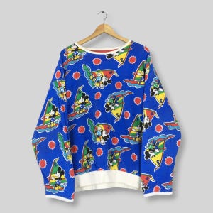 May include: A long-sleeve blue sweatshirt featuring an all-over repeating print of Mickey Mouse windsurfing on colorful boards. The design includes red sun icons and yellow, green, and red sails. The garment has a white ribbed hem and cuffs.