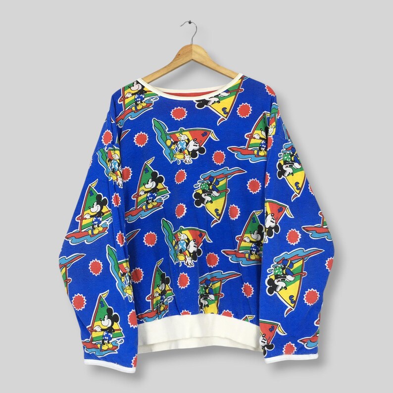 May include: A long-sleeve blue sweatshirt featuring an all-over repeating print of Mickey Mouse windsurfing on colorful boards. The design includes red sun icons and yellow, green, and red sails. The garment has a white ribbed hem and cuffs.