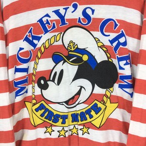 May include: A long-sleeve shirt with horizontal red and white stripes featuring a graphic of Mickey Mouse wearing a sailor hat. The text reads MICKEY'S CREW in blue and FIRST MATE in blue on a yellow banner, with four yellow stars below the banner.