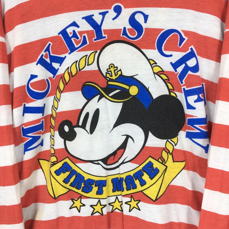 May include: A long-sleeve shirt with horizontal red and white stripes featuring a graphic of Mickey Mouse wearing a sailor hat. The text reads MICKEY'S CREW in blue and FIRST MATE in blue on a yellow banner, with four yellow stars below the banner.
