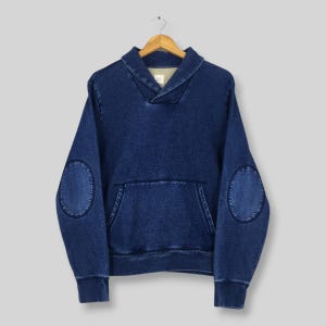 May include: A blue denim sweatshirt with a shawl collar and a kangaroo pocket. The long-sleeved top features elbow patches and is hanging on a wooden hanger. The GAP logo is visible on the collar.