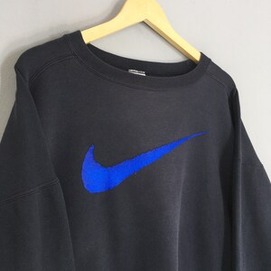 Op de afbeelding: Zwart sweatshirt met ronde hals en een groot blauw Nike-logo op de voorkant. Het Nike-logo is een opvallend element. Het sweatshirt hangt aan een houten kleerhanger tegen een lichtgrijze achtergrond.