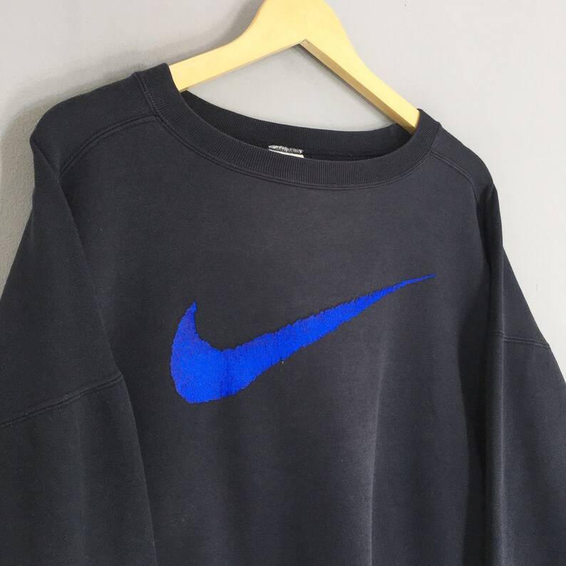 Op de afbeelding: Zwart sweatshirt met ronde hals en een groot blauw Nike-logo op de voorkant. Het Nike-logo is een opvallend element. Het sweatshirt hangt aan een houten kleerhanger tegen een lichtgrijze achtergrond.