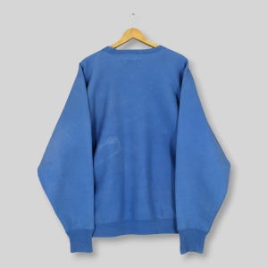 May include: A light blue sweatshirt with a crew neck and long sleeves. The sweatshirt is hanging on a wooden hanger against a light grey background. The cuffs and hem are ribbed. There is a small stain on the front of the sweatshirt.