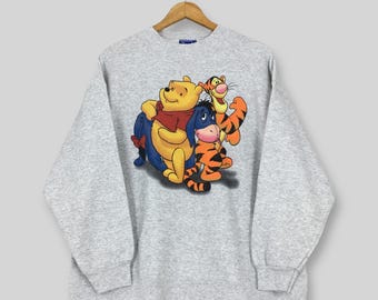 May include: A light gray sweatshirt featuring a colorful graphic of Winnie the Pooh, Tigger, and Eeyore. The cartoon characters are in a group hug. The sweatshirt has long sleeves and a crew neck.