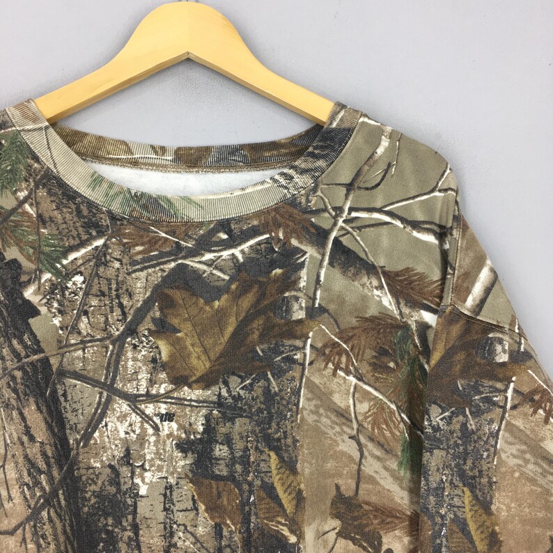 Op de afbeelding: Een sweatshirt met camouflagepatroon hangt aan een houten hanger. De stof heeft een realistische print van bomen, takken en bladeren in bruin-, groen- en beigetinten. De ronde hals en mouwen zijn zichtbaar.