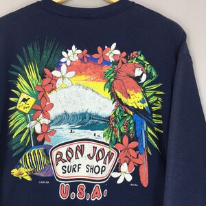May include: Dark blue sweatshirt with a vibrant print of a parrot, tropical flowers, and a wave scene. The design includes the text "Ron Jon Surf Shop U.S.A." and a yellow sign with a kangaroo. A colourful, eye-catching design.