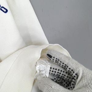 May include: Close-up of a white knit sweater with a blue embroidered detail. The sweater's texture is visible, and a portion of the sleeve is turned inside out, revealing the interior. The image is well-lit, highlighting the fabric's details.