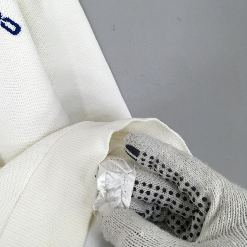 May include: Close-up of a white knit sweater with a blue embroidered detail. The sweater's texture is visible, and a portion of the sleeve is turned inside out, revealing the interior. The image is well-lit, highlighting the fabric's details.