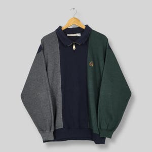 May include: A color-blocked sweatshirt with a collared neckline and a quarter-zip closure. The sweatshirt features panels of gray, navy, and green. A small embroidered emblem is on the green panel. The garment is hanging on a wooden hanger.