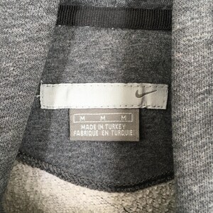 Może przedstawiać: Zbliżenie szarej metki bluzy z kapturem Nike. Etykieta zawiera logo Nike swoosh oraz napis "MADE IN TURKEY FABRIQUE EN TURQUIE". Etykieta ma r&oacute;wnież trzy wskaźniki rozmiaru "M".