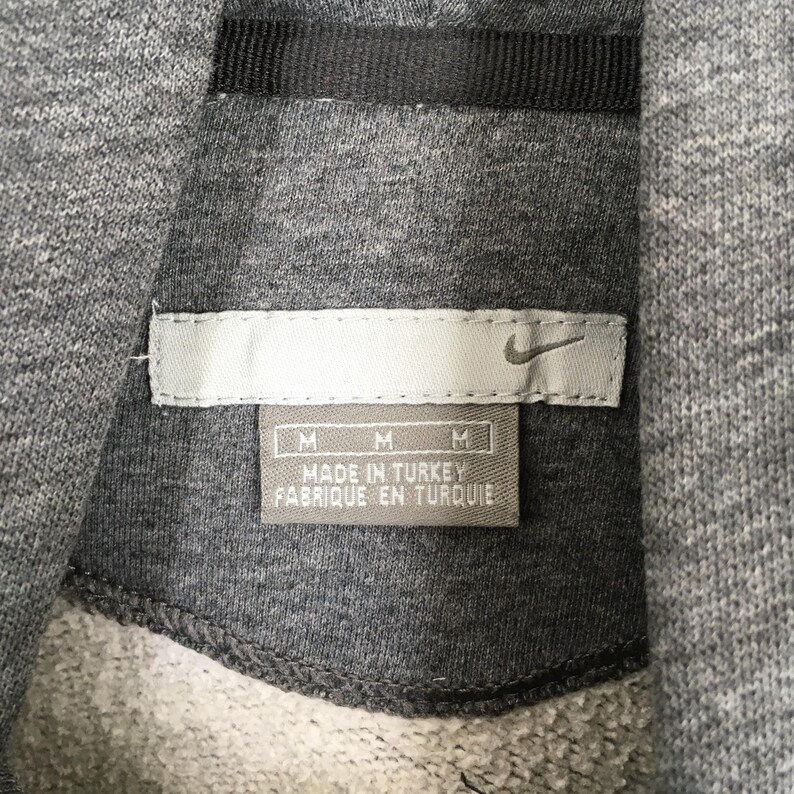 Może przedstawiać: Zbliżenie szarej metki bluzy z kapturem Nike. Etykieta zawiera logo Nike swoosh oraz napis "MADE IN TURKEY FABRIQUE EN TURQUIE". Etykieta ma r&oacute;wnież trzy wskaźniki rozmiaru "M".