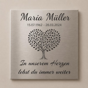 Könnte beinhalten: Eine quadratische Gedenktafel aus gebürstetem silberfarbenem Metall mit der Gravur Maria Müller, den Daten 15.07.1962 - 28.03.2024, einem Baum mit herzförmiger Krone sowie dem Text In unserem Herzen lebst du immer weiter.