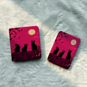 May include: Two rectangular e-reader cases featuring a vibrant magenta background with a graphic illustration of three black cats sitting on a ledge under a full moon and cherry blossom branches. The design includes dark pink floral accents.