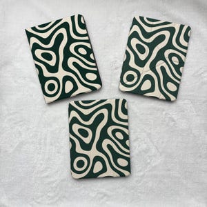May include: Three rectangular tablet cases featuring a dark forest green and cream-colored abstract wavy pattern. The designs consist of fluid, organic shapes that create a continuous, interconnected look across the surfaces of the three covers.
