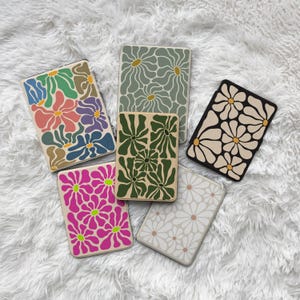May include: A collection of six rectangular cases with floral designs. The cases feature various color schemes, including green, pink, and neutral tones. The floral patterns are stylized and modern, with a retro aesthetic.