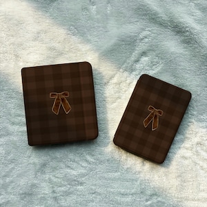 May include: Two rectangular cases featuring a dark brown and black plaid pattern. Each case is centered with a small, brown velvet-textured bow embellishment. The items are positioned side by side, displaying a consistent design and color scheme.