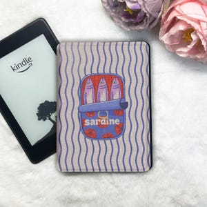 Puede incluir: Funda Kindle con diseño de lata de sardinas. La funda presenta una ilustración de dibujos animados de tres sardinas rosas en una lata azul con la palabra "sardina" en rojo. El fondo tiene rayas azules verticales sobre un fondo gris claro.