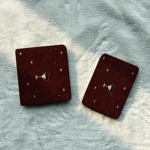 May include: Two dark red, velvet-textured notebooks featuring a gold-colored graphic of a black cat sitting inside a martini glass, surrounded by small, scattered gold-colored star icons across the front covers of both items.