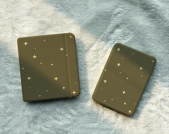 May include: Two olive green rectangular electronic reading devices with rounded corners, each featuring a scattered pattern of small, four-pointed gold stars across the front covers. The devices are positioned side by side.