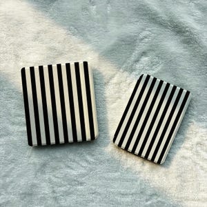 Puede incluir: Dos espejos compactos rectangulares a rayas blancas y negras. Los espejos tienen un acabado brillante y están colocados sobre una superficie suave y blanca texturizada. Las rayas son verticales en un espejo y diagonales en el otro.