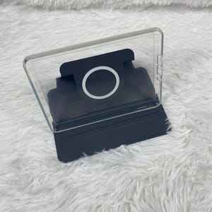 May include: A dark grey tablet case featuring a clear back panel and a built-in circular magnetic charging ring. The case includes a foldable stand mechanism and precise cutouts along the side for buttons, providing a secure fit for a device.