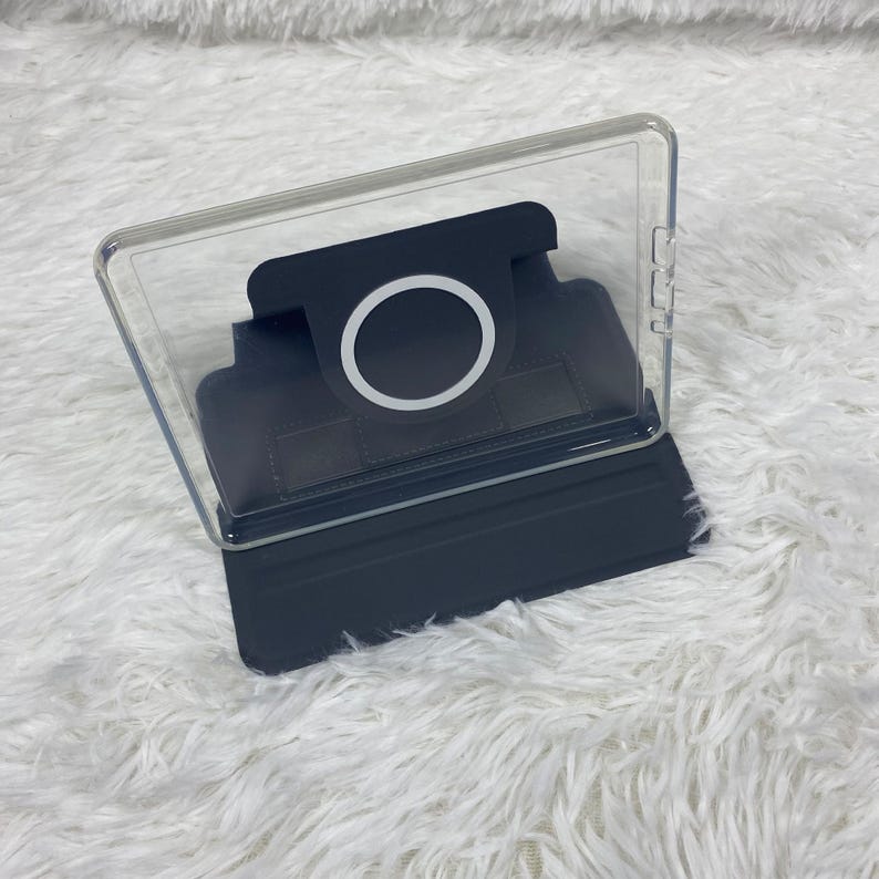 May include: A dark grey tablet case featuring a clear back panel and a built-in circular magnetic charging ring. The case includes a foldable stand mechanism and precise cutouts along the side for buttons, providing a secure fit for a device.