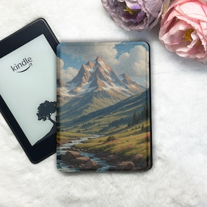 Puede incluir: Un lector de libros electrónicos Kindle junto a una funda con una pintura de paisaje de una cordillera, un río y un valle. La pantalla del Kindle muestra la palabra "kindle" y una silueta de árbol. La funda tiene un borde negro.